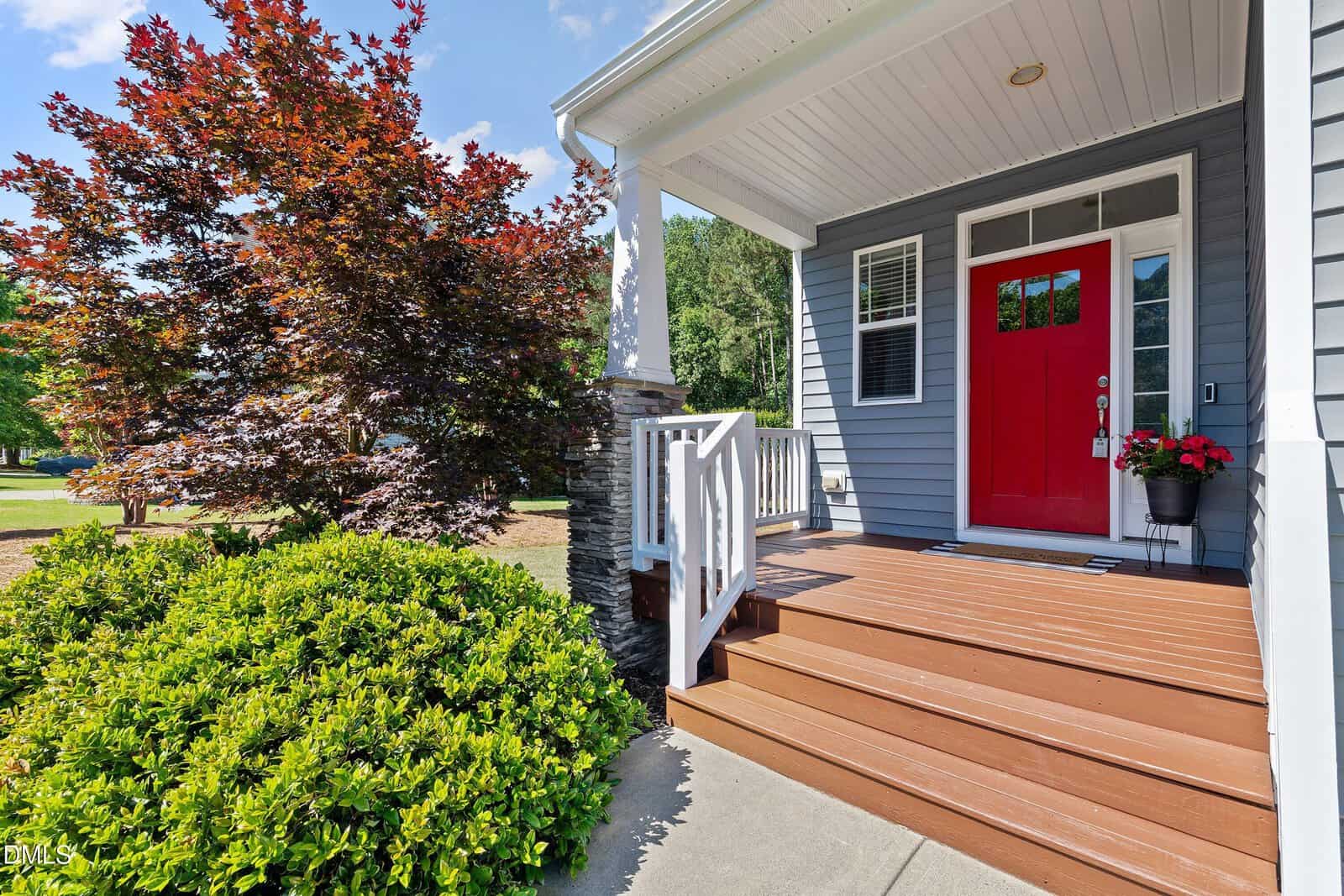 A wooden front porch with a bright red door, potted flowers, and white railing, surrounded by green shrubs and a red-leafed tree under a blue sky.