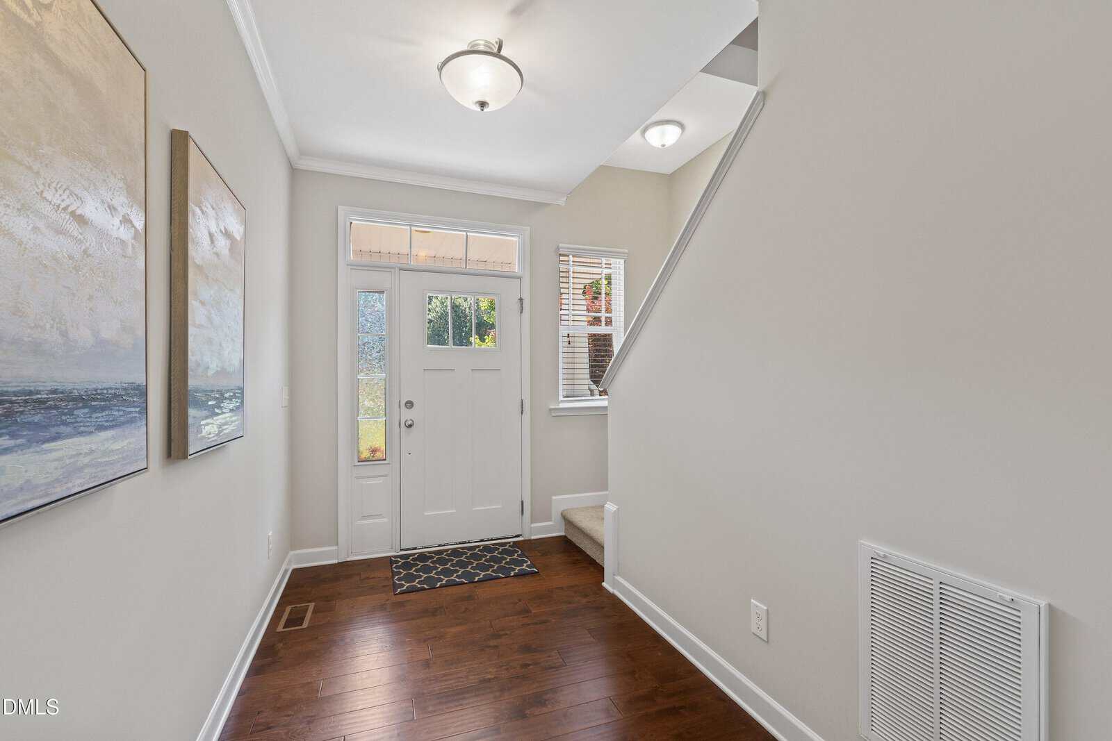 A bright entryway with a white front door, sidelights, hardwood floor, beige walls, two wall paintings, a stairway with carpet, and a small window near the stairs.