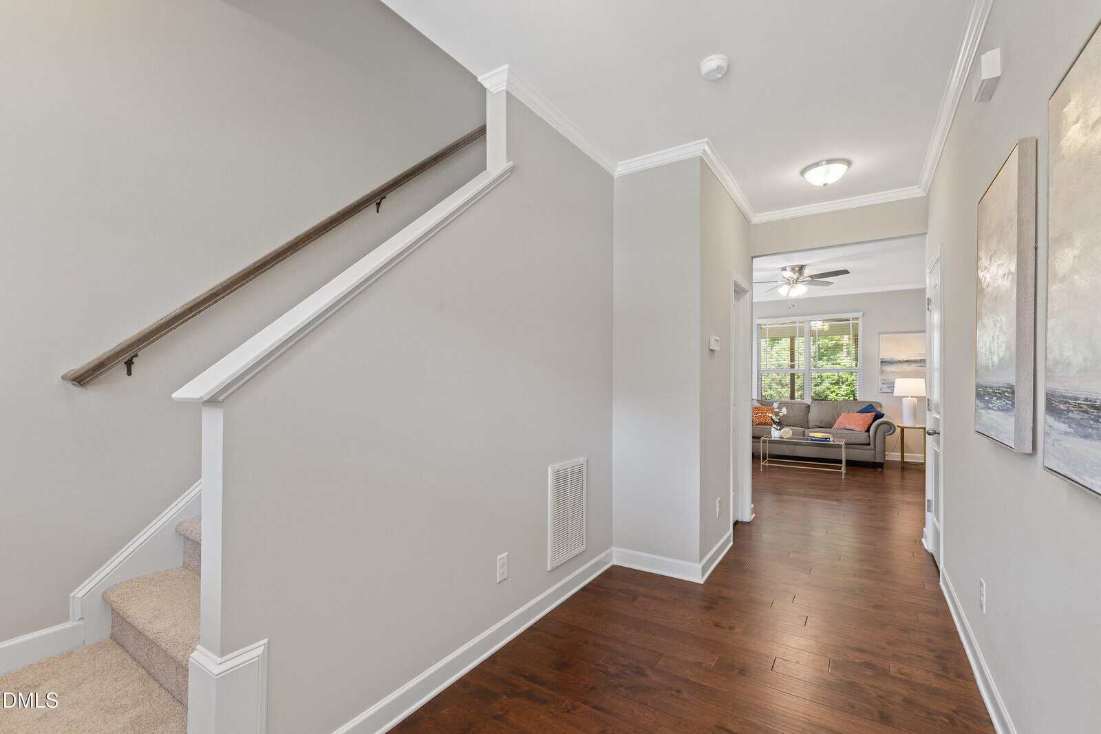Bright hallway with hardwood floors, light gray walls, and white trim. Carpeted stairs are on the left. A living room with a ceiling fan and large window is visible at the end of the hall.