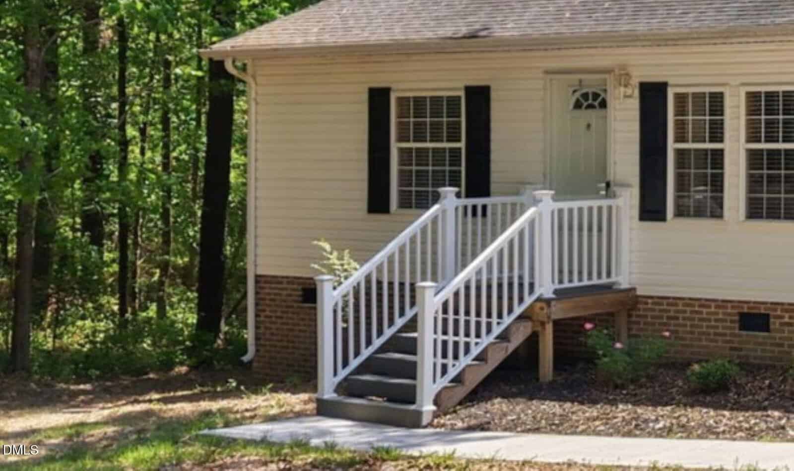 A small front porch with white railings and steps leads to a light green door on a beige house with black shutters, surrounded by trees and greenery.