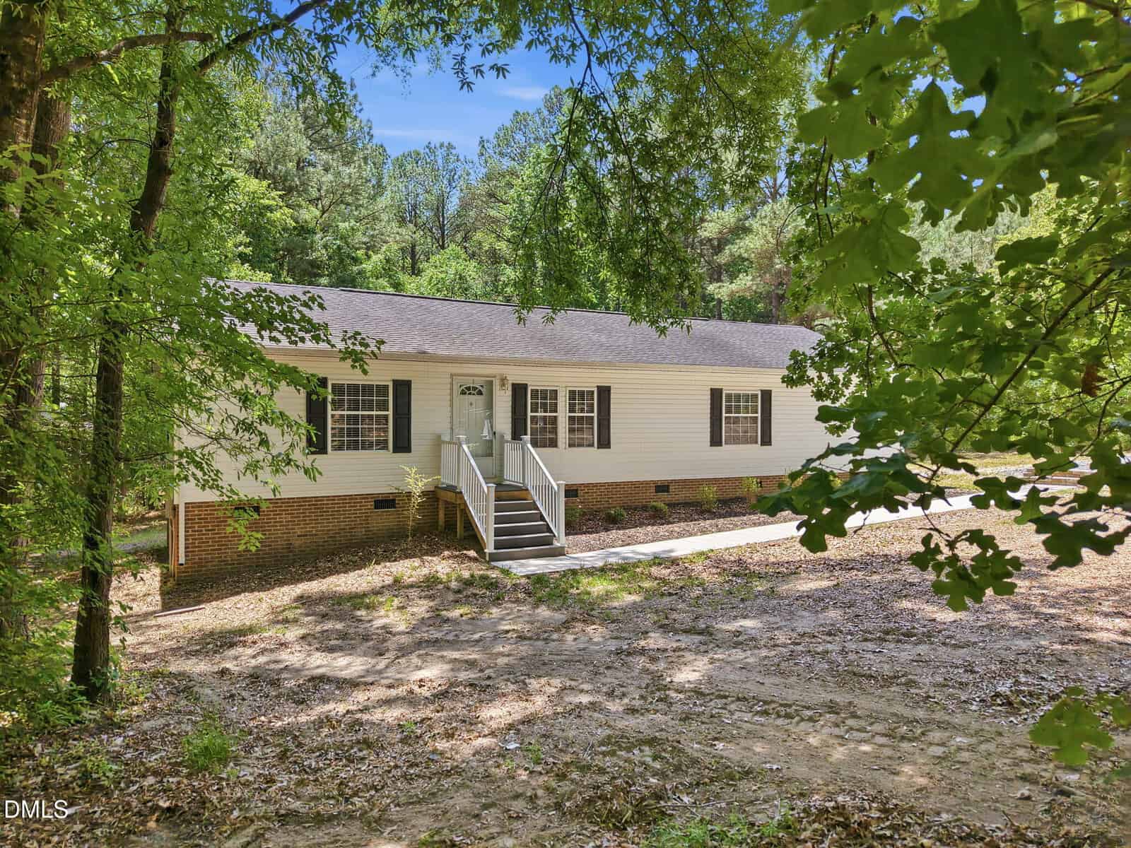 A single-story white house with a brick foundation, front steps, and a small porch, surrounded by trees and greenery under a clear blue sky.