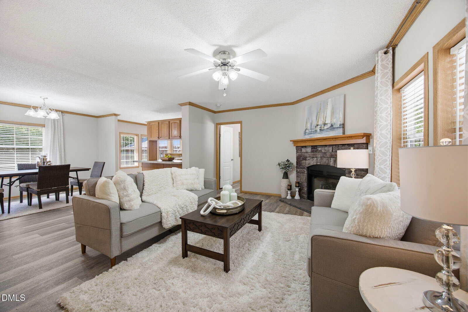 A bright living room features two sofas, a wooden coffee table, a fireplace with a painting above it, light walls, wood trim, and large windows. The dining area and kitchen are visible in the background.