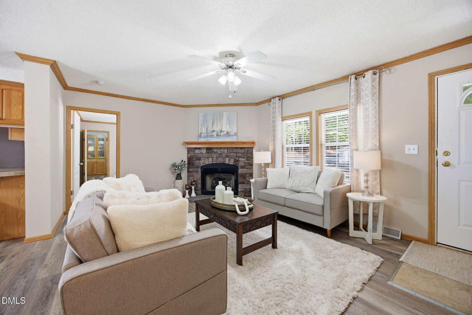 A bright living room features neutral furnishings, a white rug, a stone fireplace, two sofas, a coffee table, and large windows with curtains. A ceiling fan and decor create a cozy, inviting atmosphere.