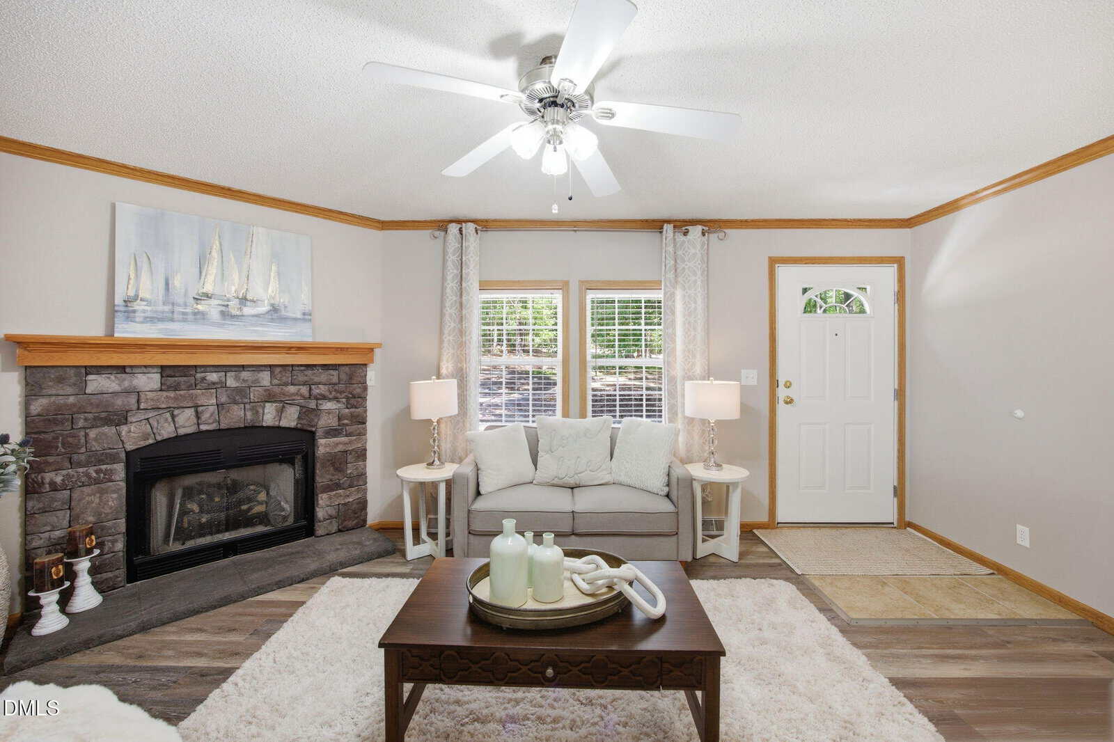 A cozy living room with a stone fireplace, sailboat art, gray couch, two side tables with lamps, a wooden coffee table, and a ceiling fan, all in neutral tones with wood trim and light decor.