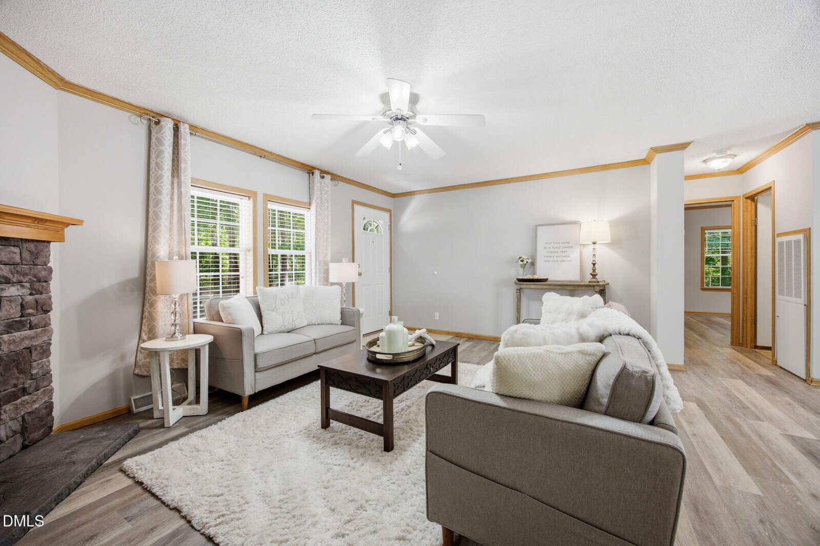 Bright living room with gray sofas, white rug, coffee table, and fireplace. Light wood floors, white walls, large windows with curtains, ceiling fan, and decorative accents in a cozy, inviting arrangement.