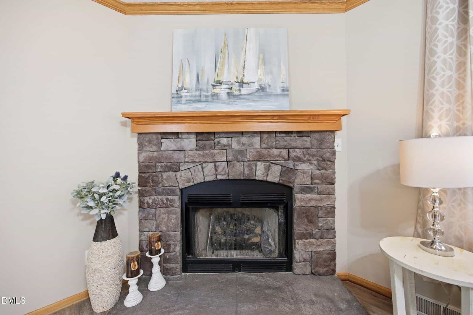 A stone fireplace with a wooden mantle, sailboat painting above, and decorative items including a vase with greenery, two candles, and a table lamp on the side.