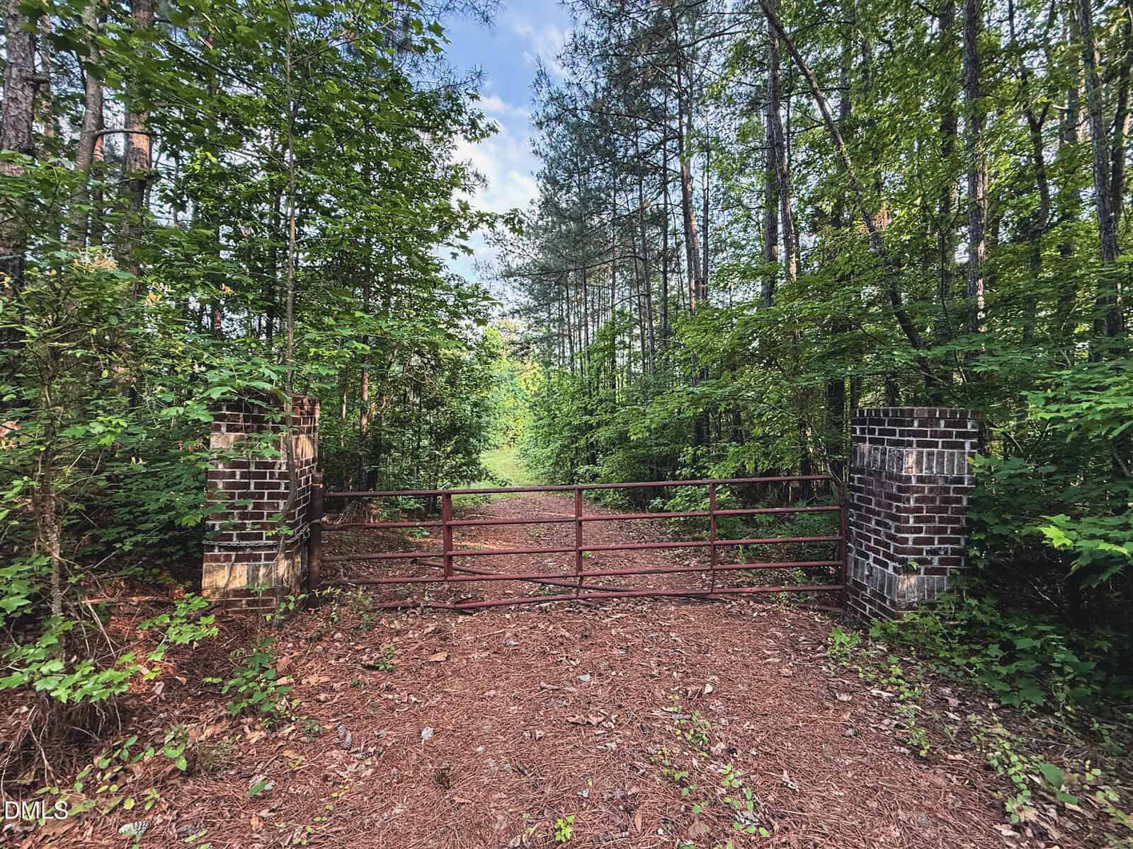A rust-colored metal gate stands between two weathered brick pillars, blocking a dirt path surrounded by dense green trees and foliage under a partly cloudy sky.