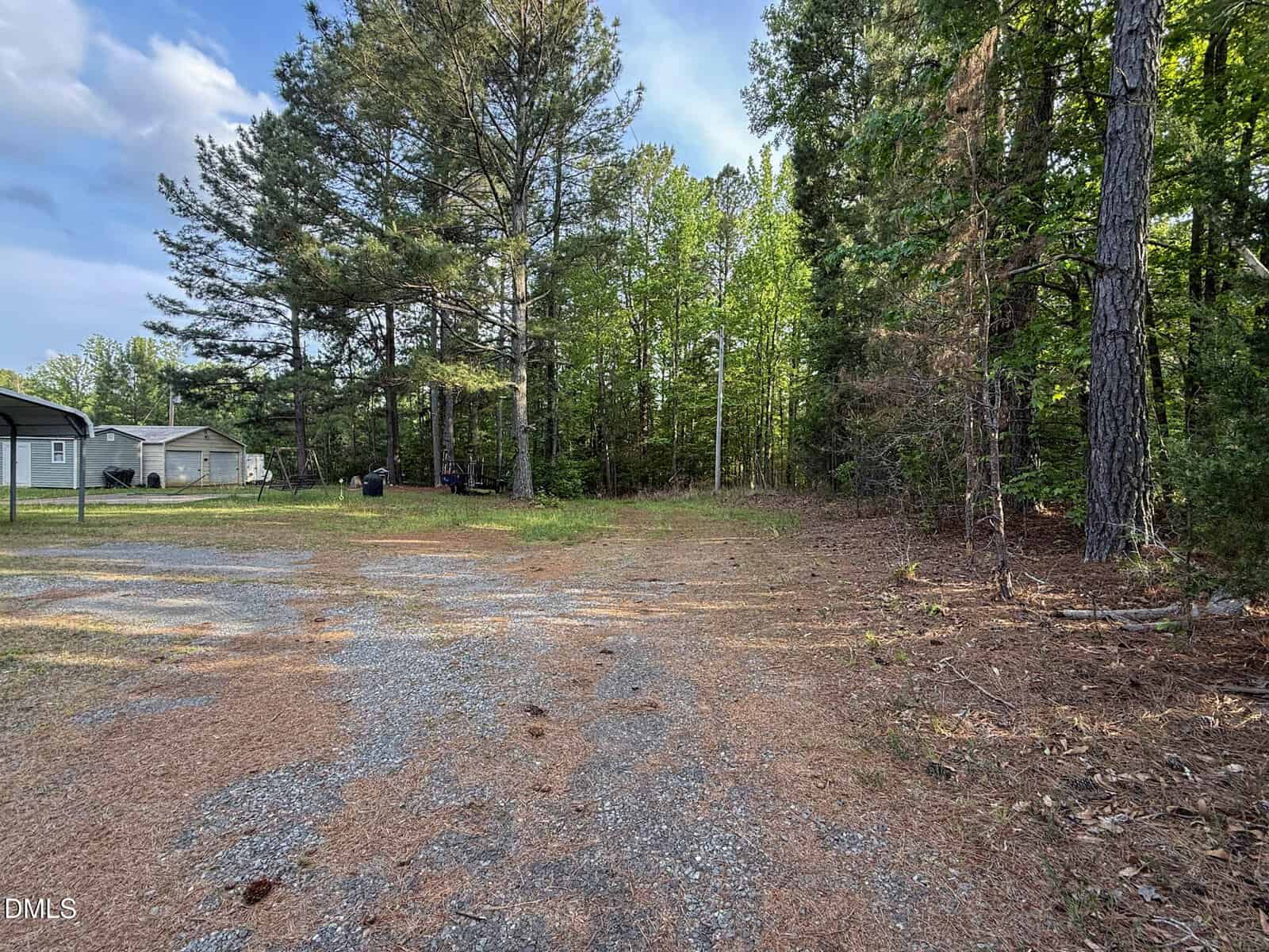 Gravel driveway leading to a wooded area with tall trees; a small house and shed are visible on the left under a partly cloudy sky.