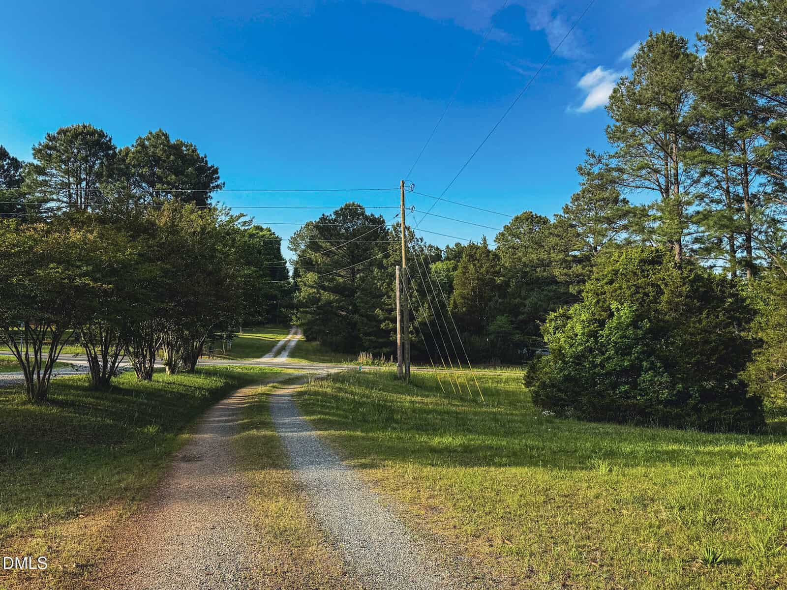 A gravel driveway curves through a grassy area with trees on both sides, leading to a paved road in the distance under a blue sky with scattered clouds. Power lines run alongside the driveway.