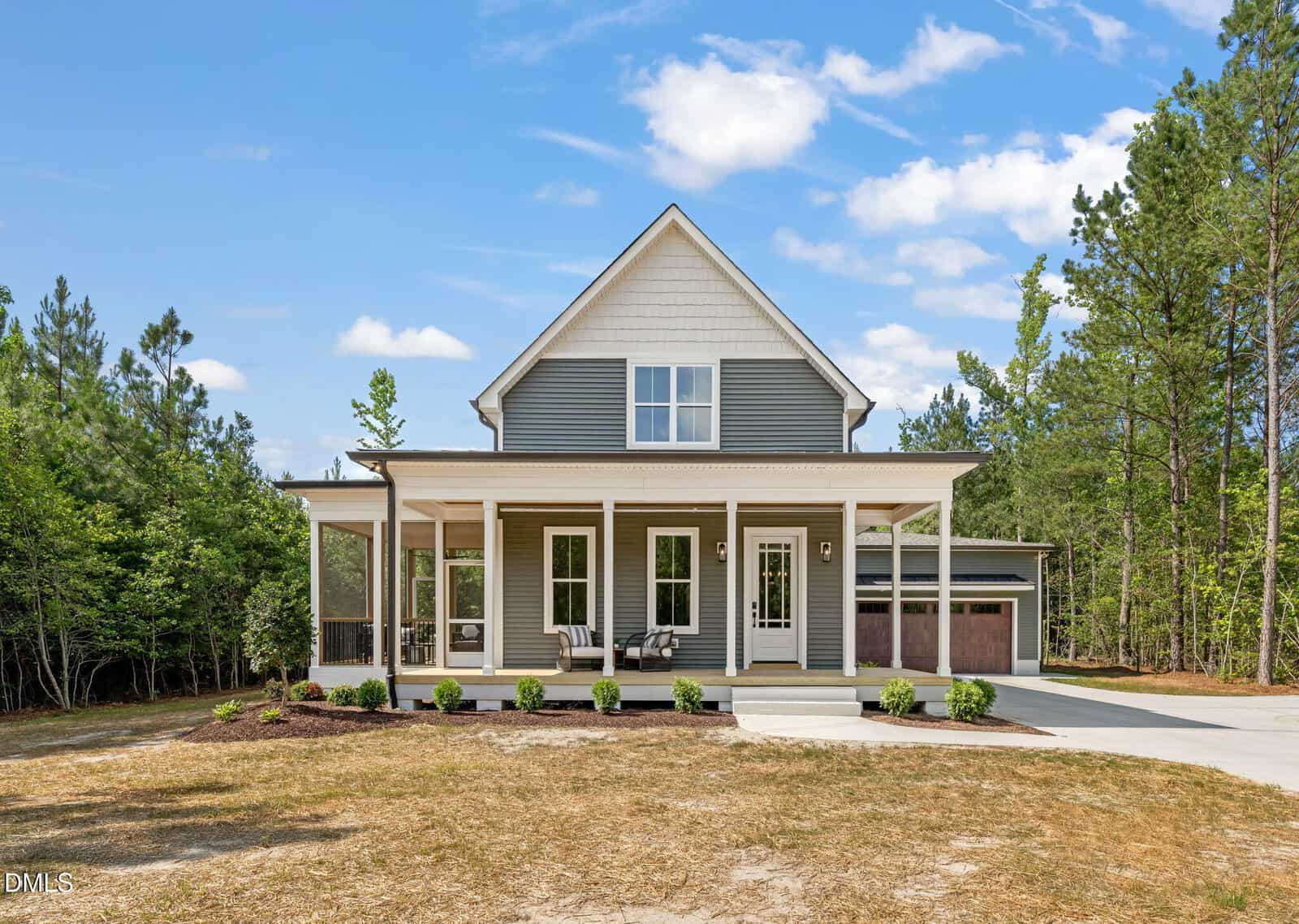 A modern two-story house with gray siding, a covered front porch, large windows, and a double garage, surrounded by trees and a partly cloudy blue sky.
