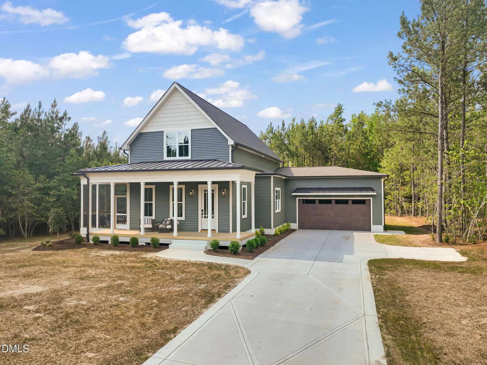 A modern gray house with a large porch, white trim, and a two-car garage sits on a spacious lot with trees in the background and a concrete driveway leading to the front entrance.