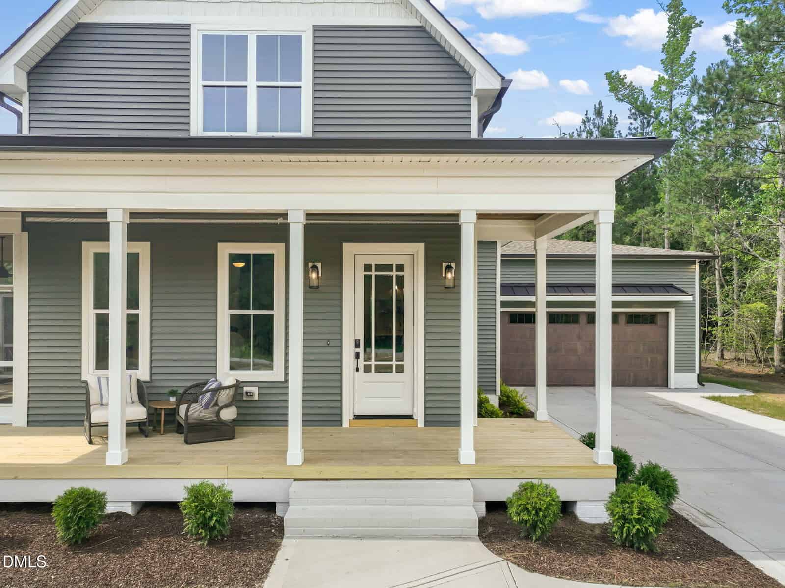 A modern house with gray siding, a white-trimmed porch, two chairs with a small table, a white front door, and neatly landscaped bushes in front. A detached garage and trees are in the background.