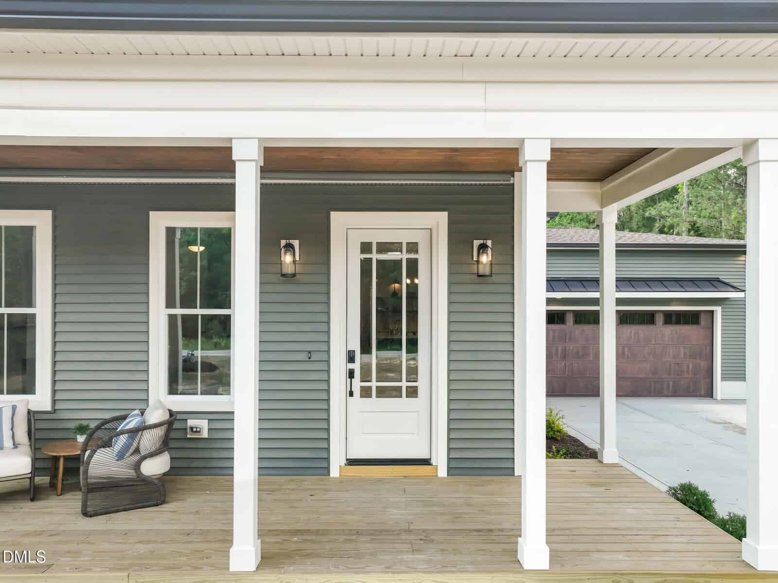 A modern house front porch with white columns, a glass-paneled door, two wall lanterns, greenish-gray siding, and a seating area. A driveway leads to a detached garage with wooden doors in the background.