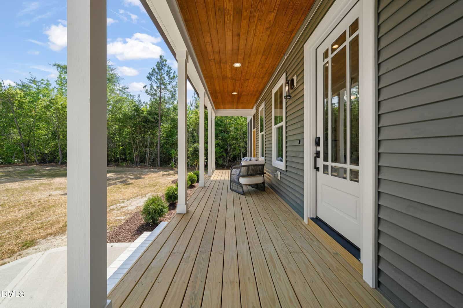 A spacious, covered front porch with wooden flooring, white columns, gray siding, a white door, and outdoor seating. The porch overlooks a grassy yard bordered by trees under a blue sky.