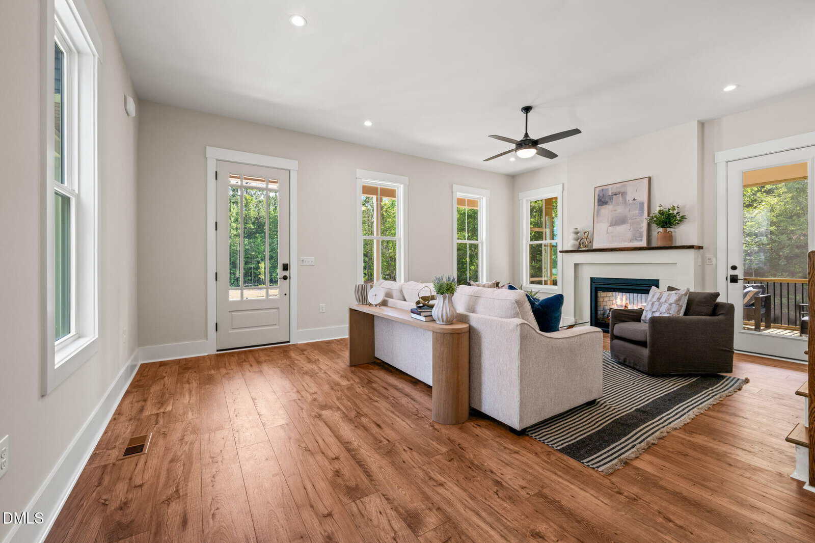 Bright living room with wood floors, neutral walls, large windows, a gray sofa, armchair, and a fireplace. Glass doors lead to a deck with views of greenery outside. Modern decor with natural light filling the space.