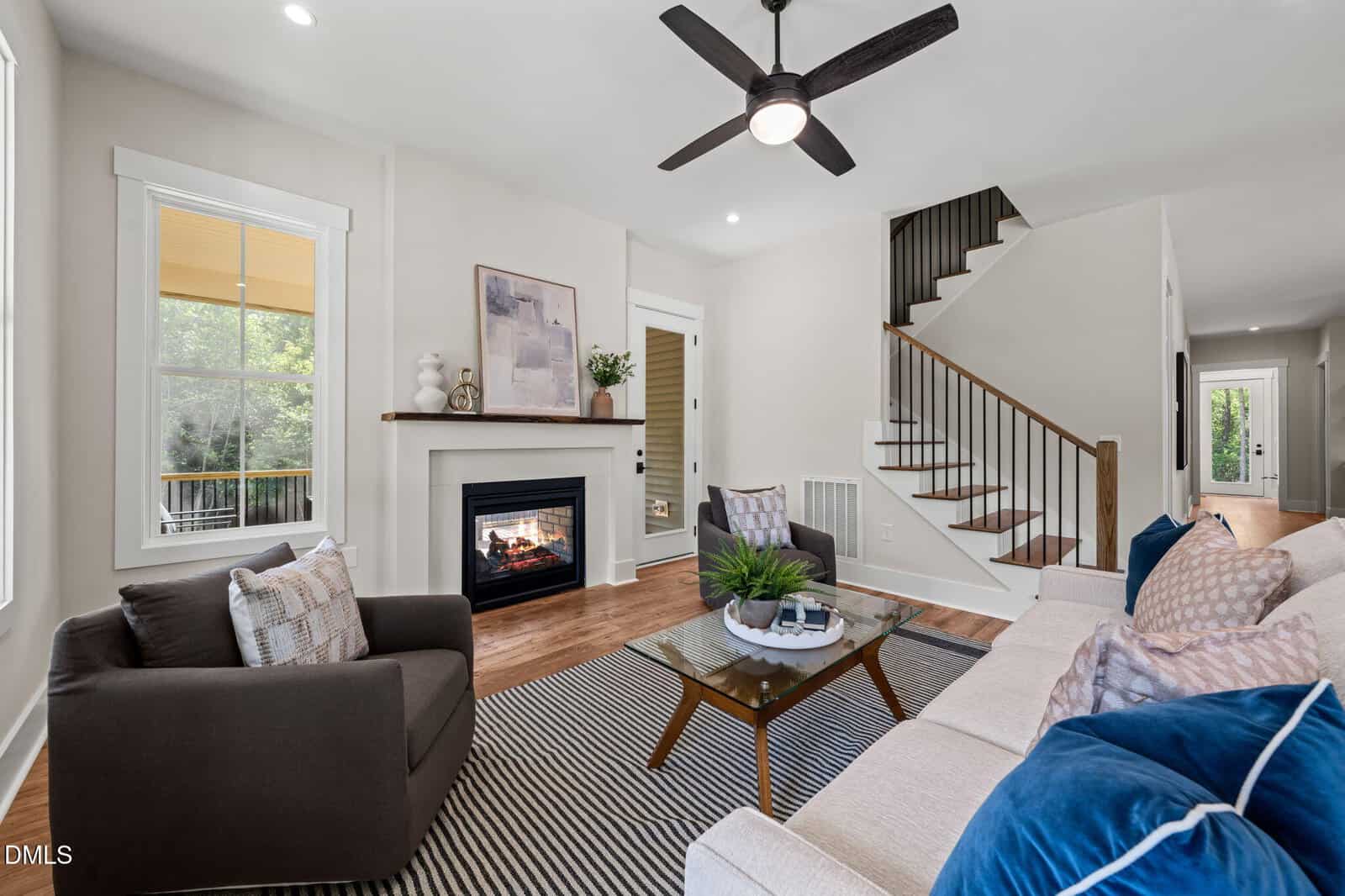 Modern living room with a beige sofa, brown armchair, glass coffee table, fireplace, striped rug, wall art, and a ceiling fan. Stairs lead up, and large windows let in natural light.