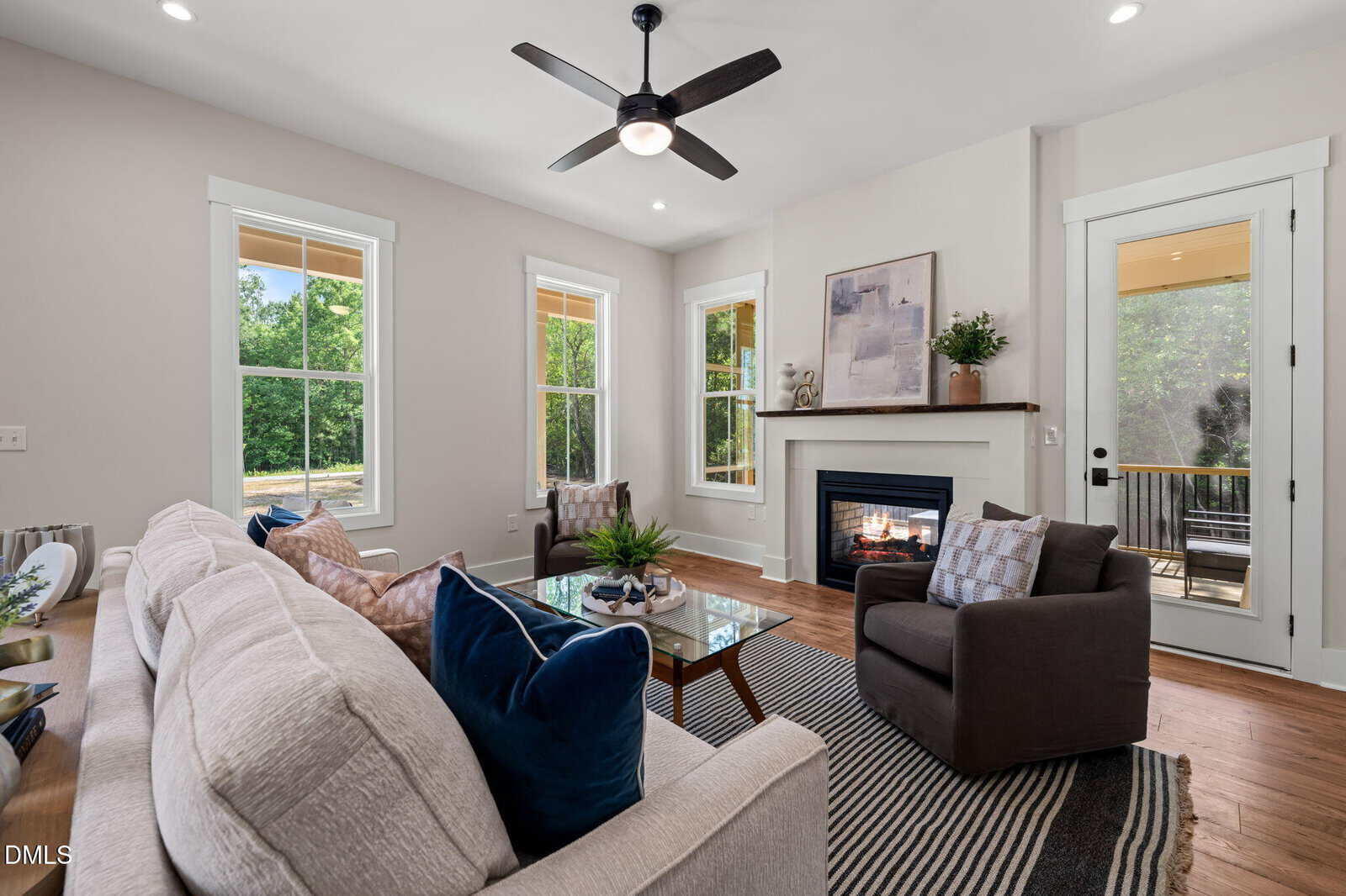 Bright living room with a beige sofa, blue and brown pillows, armchair, gas fireplace, artwork, ceiling fan, large windows, glass door, and a view of greenery outside. Modern decor with cozy touches.