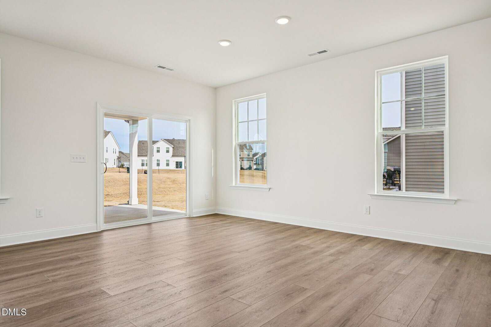 Bright, empty room with light wood flooring, white walls, two tall windows, and sliding glass doors opening to a backyard with neighboring houses visible outside.