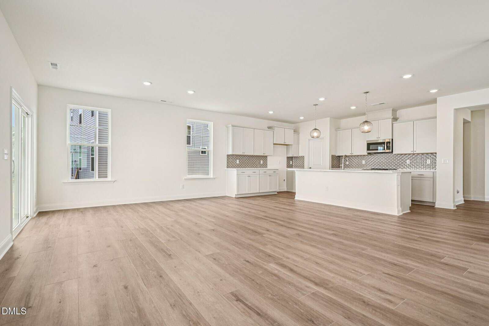 Open-concept living area with light wood flooring, white walls, large windows, and a modern kitchen featuring white cabinets, stainless steel appliances, pendant lights, and a gray tile backsplash.