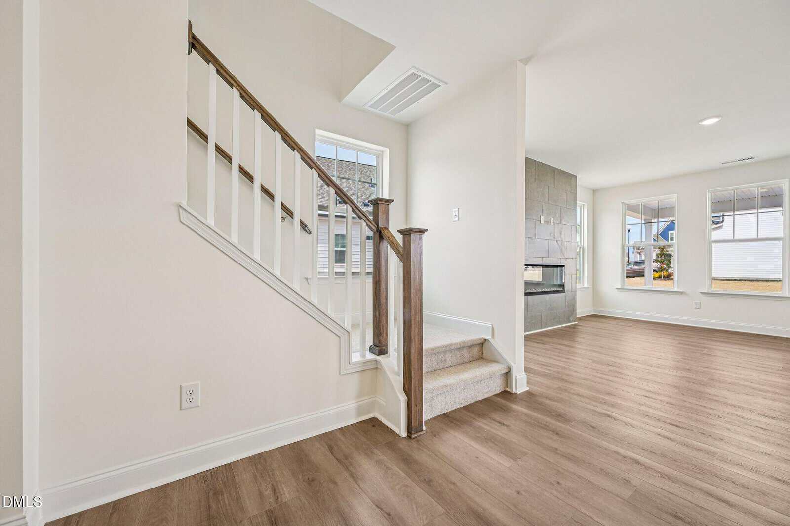 Bright, modern interior with wood flooring, white walls, a staircase with brown railing, and an open living area featuring large windows and a double-sided fireplace.