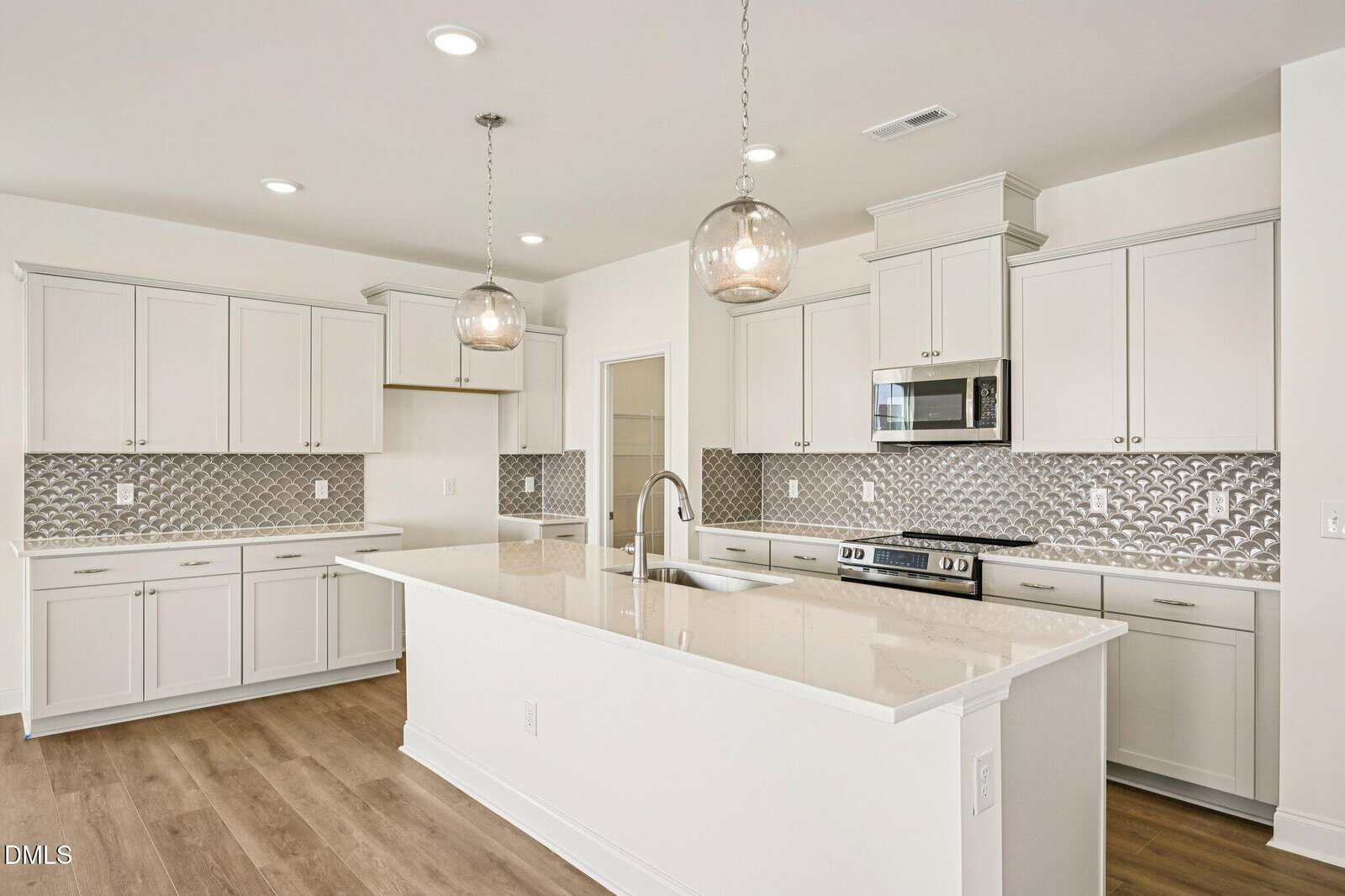 Modern kitchen with white cabinets, silver appliances, a large island with a sink, pendant lights, and patterned silver backsplash. Light wood flooring and recessed ceiling lights complete the bright, airy space.