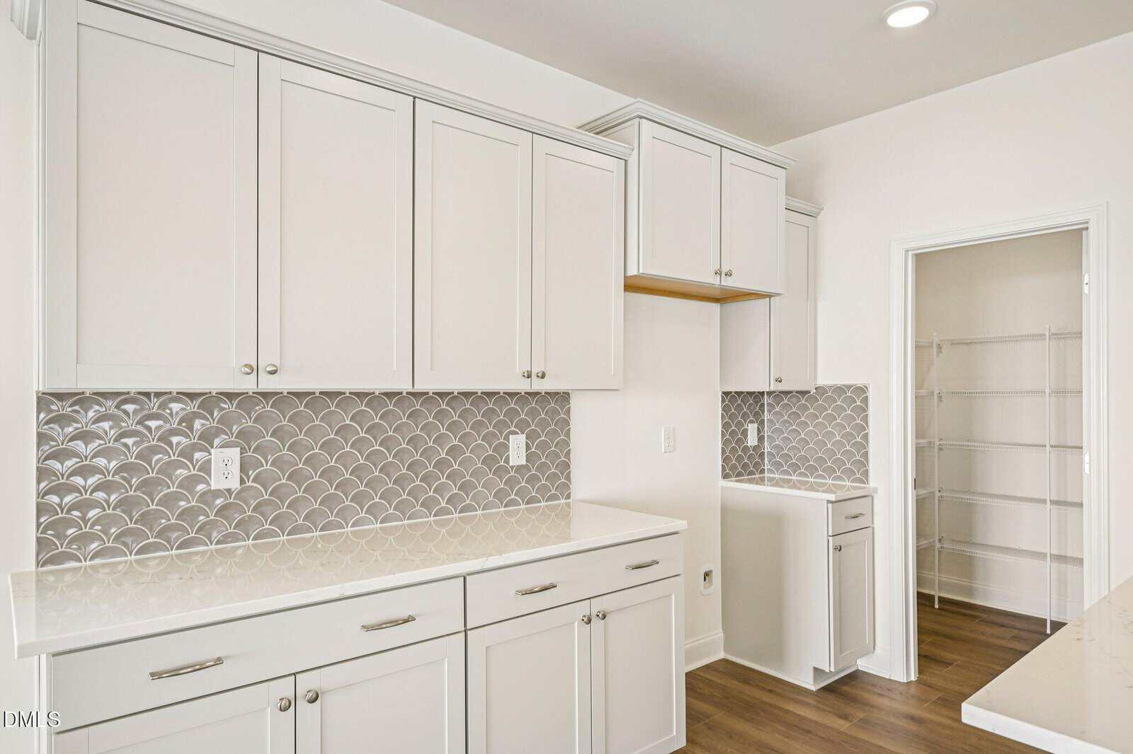 A modern laundry room with white cabinets, a patterned backsplash, wood flooring, and an open pantry with wire shelves. There are countertop spaces and multiple drawers and cabinets for storage.
