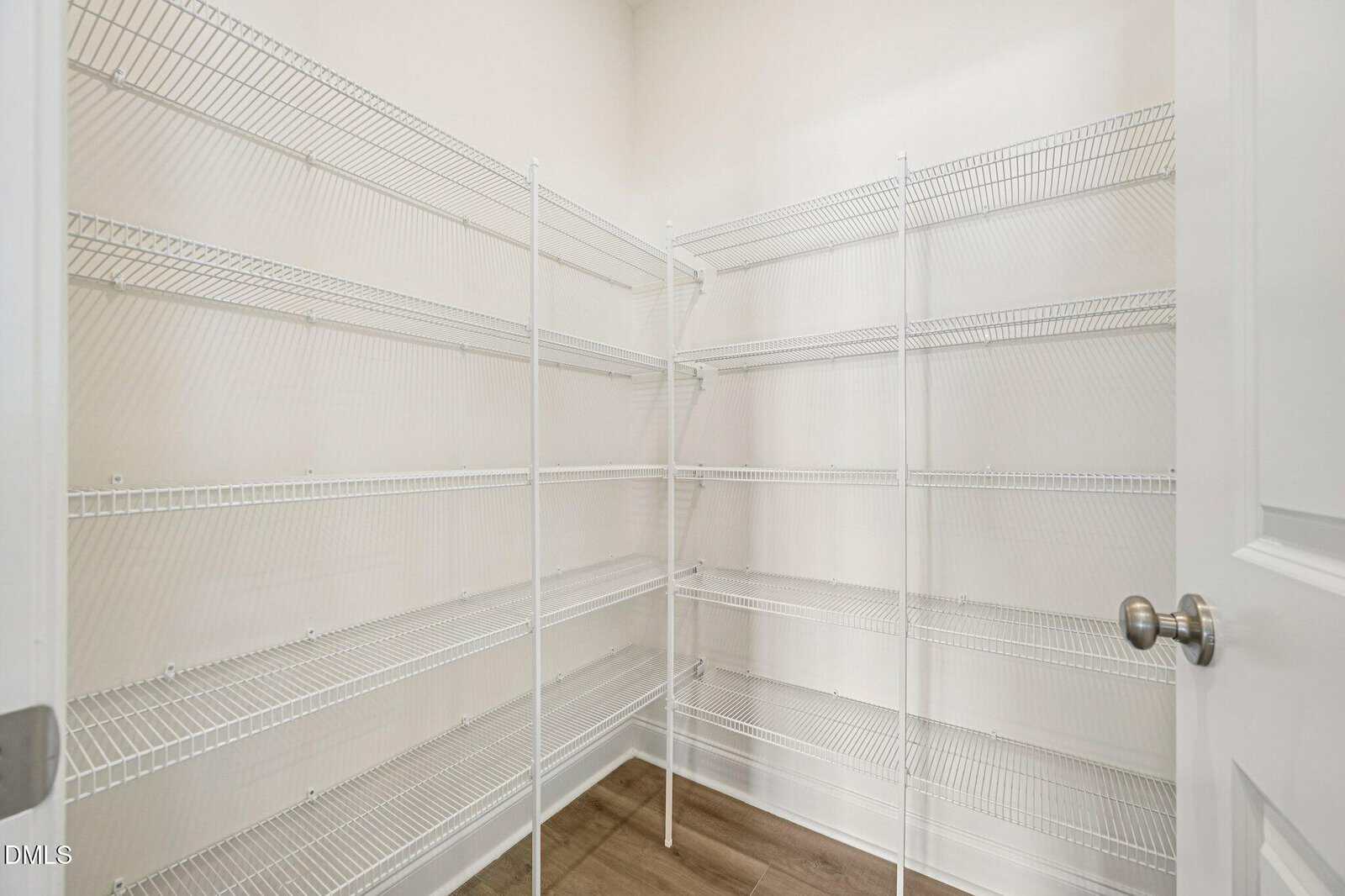 Empty walk-in pantry with white wire shelving on three walls, light-colored walls, and wood flooring. The shelves are evenly spaced and the room is well-lit with a simple white door and silver door knob visible.