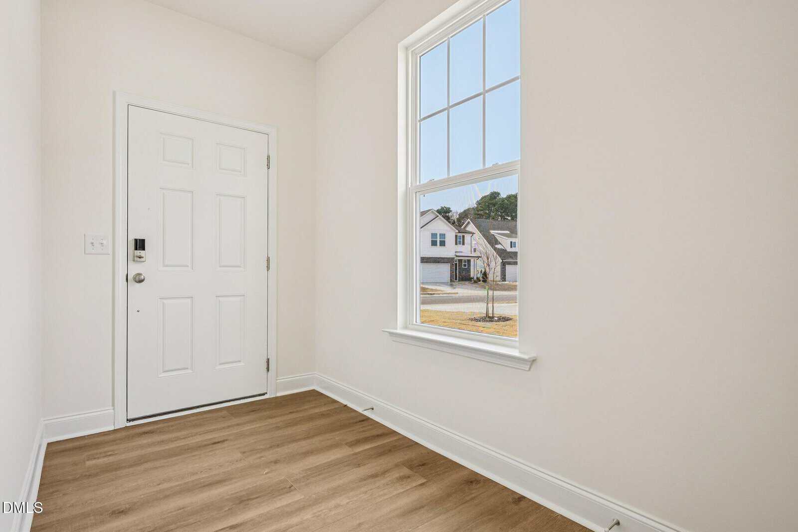 A small entryway with light-colored walls, wood flooring, a white front door with a lock, and a tall window letting in natural light, showing houses and a street outside.