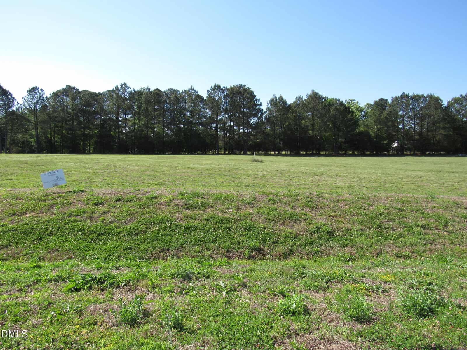 A grassy, open field with a sign on the left side, bordered by tall trees in the background under a clear blue sky.
