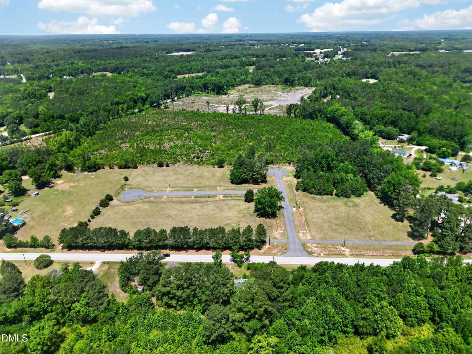 Aerial view of a rural, partially wooded landscape with open grassy areas, a curved paved road, scattered trees, and surrounding forest under a partly cloudy sky. A few buildings and vehicles are visible in the distance.