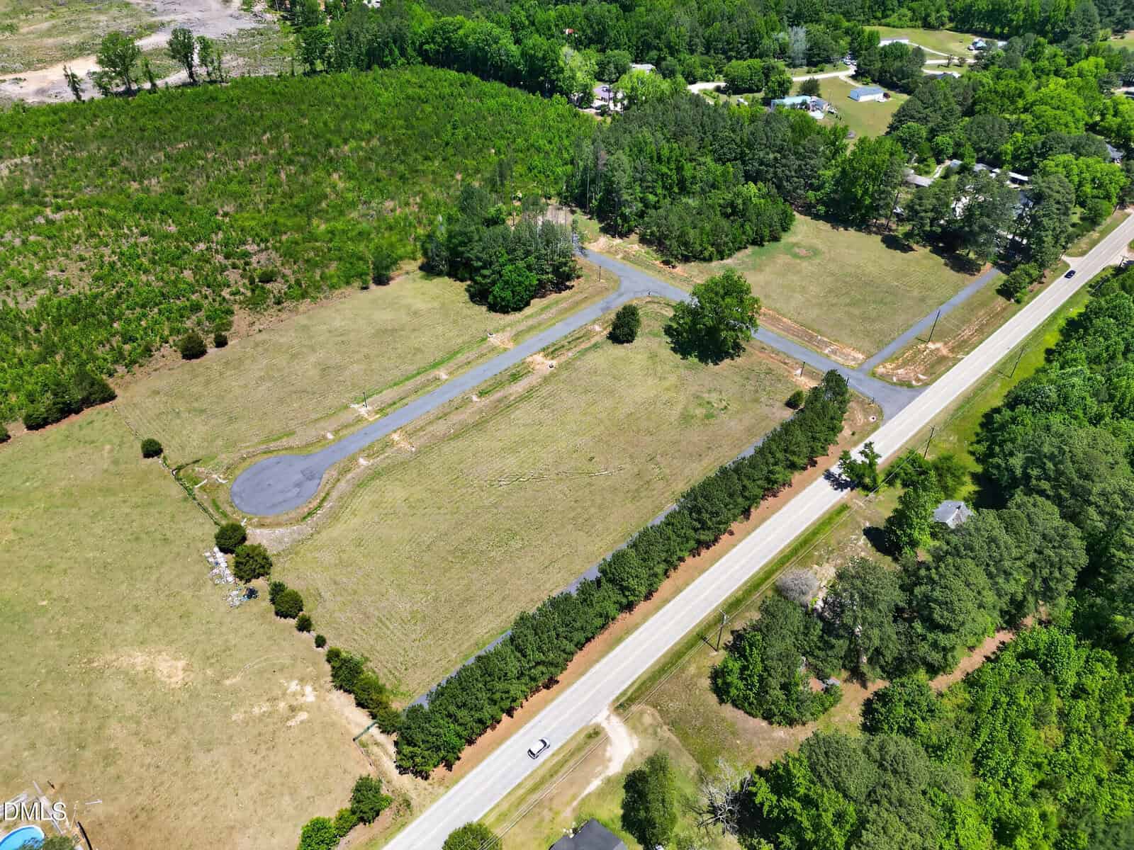 Aerial view of a rectangular, partially cleared plot of land bordered by trees and a road, with a few paved driveways and green grass areas surrounded by wooded and rural landscape.