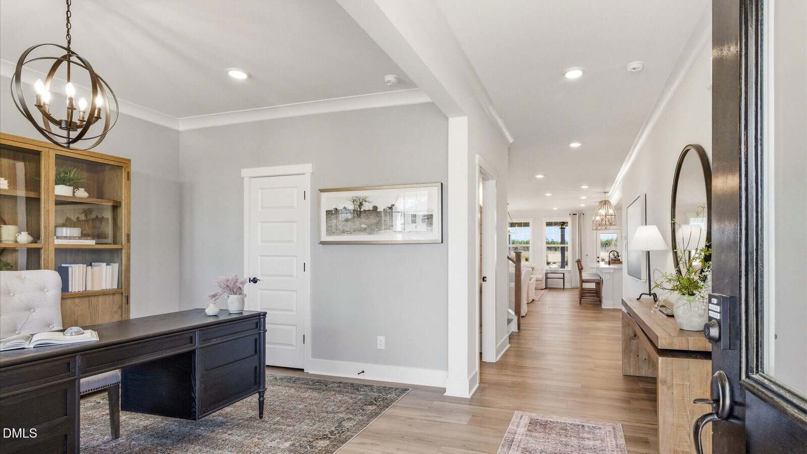 A bright home office with a black desk and shelves, light gray walls, and wood flooring, opening into a hallway that leads to a living area with large windows and modern decor.