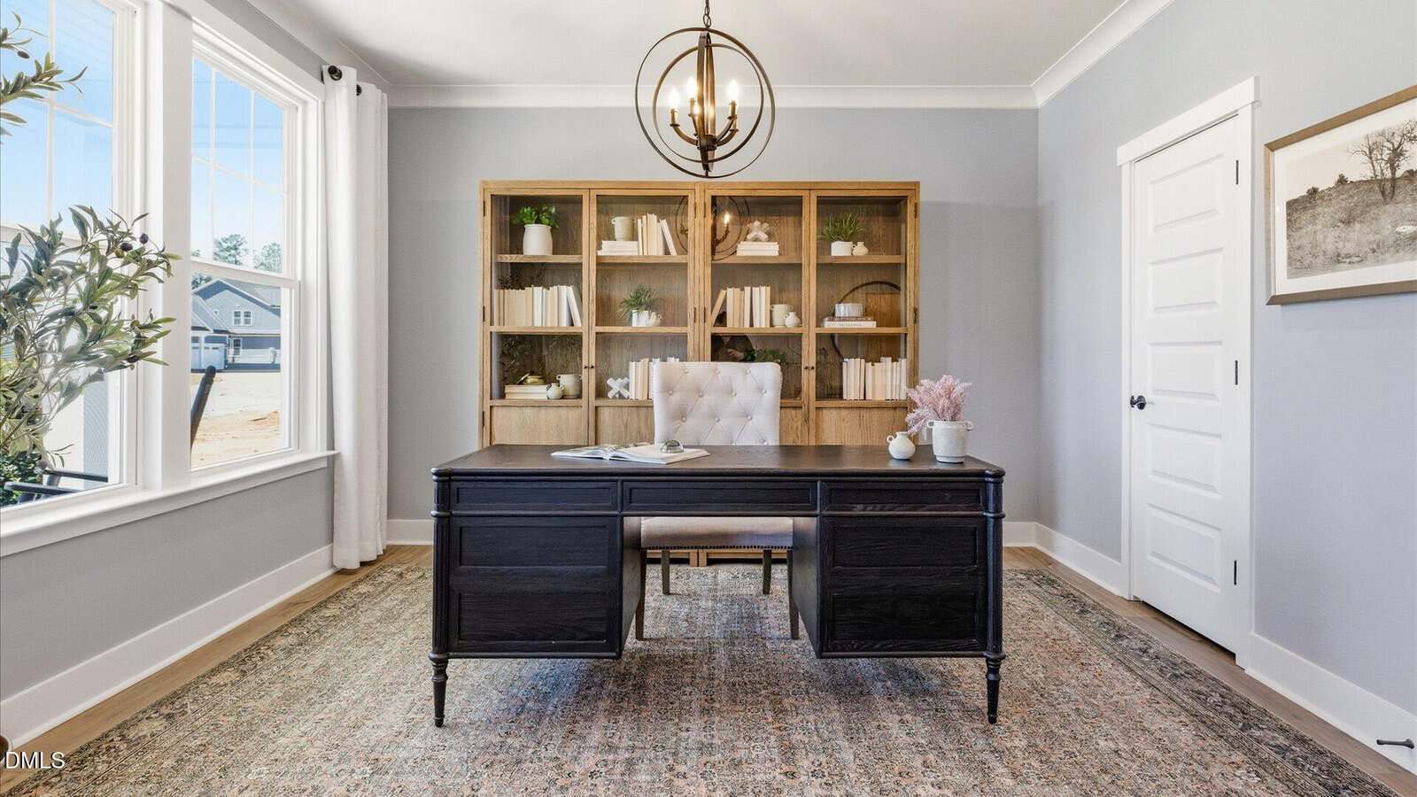 A bright home office with a dark desk, cushioned chair, bookshelf filled with books, large window, indoor plant, modern chandelier, and a gray rug on wooden floors.