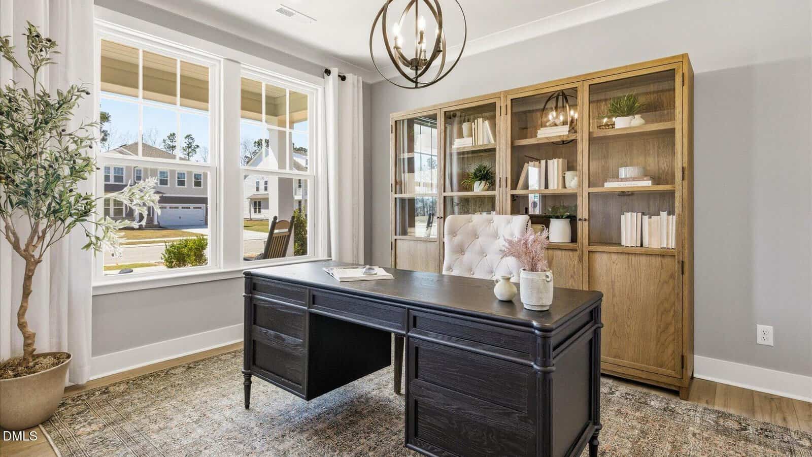 A bright home office features a large window, black desk, tufted chair, bookshelves with books and decor, a globe light fixture, and a potted plant on a patterned rug.