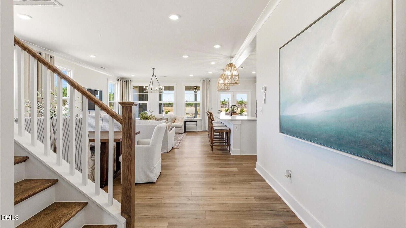 Bright, modern open-plan living space with wood floors, white walls, large windows, and a staircase on the left. Dining and sitting areas lead to a kitchen with pendant lights. A large painting hangs on the right wall.