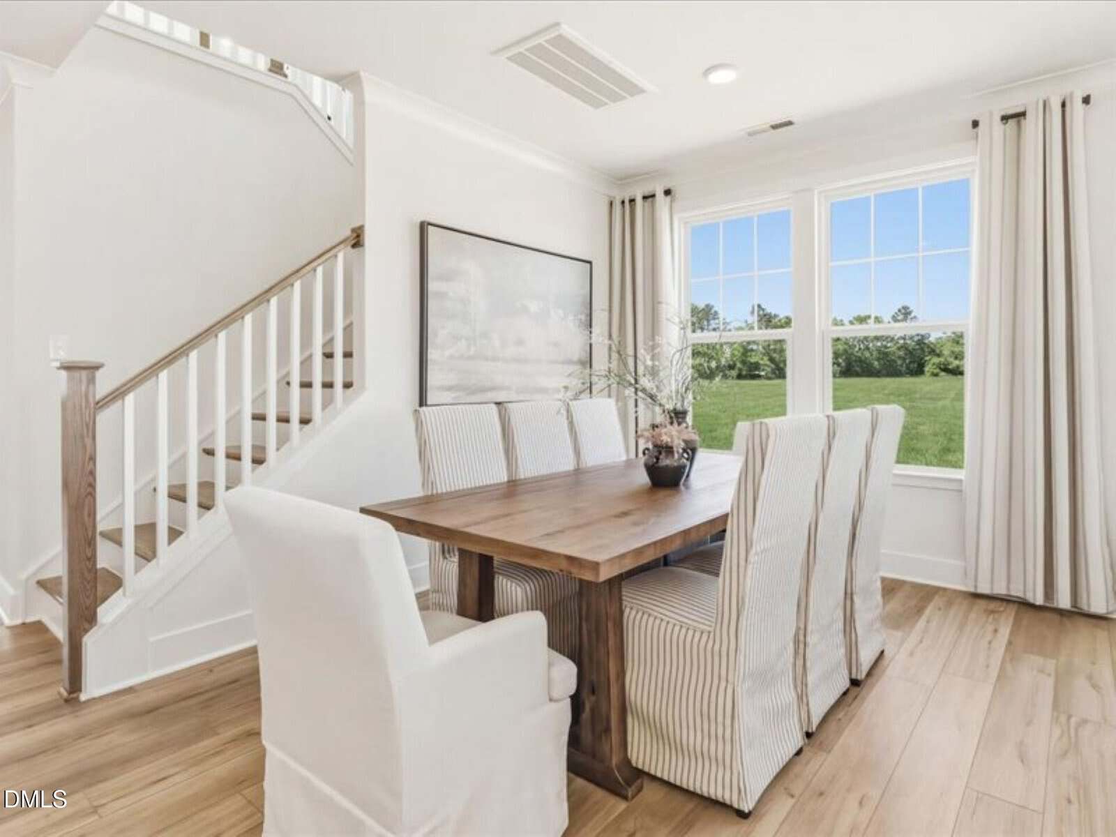 Bright dining area with a wooden table, six white upholstered chairs, large window with curtains, light wood flooring, and a staircase with white railing in the background. A framed picture hangs on the wall.