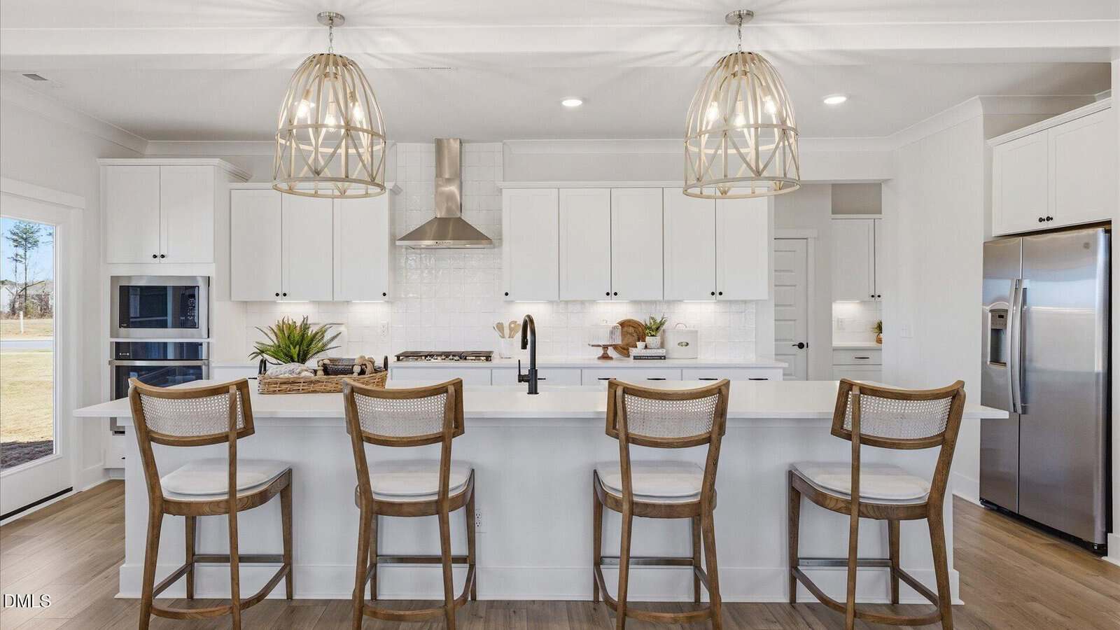 Modern kitchen with white cabinets, stainless steel appliances, four wooden barstools at a large island, wicker pendant lights above, and light wood flooring. Natural light streams in from the left side window.
