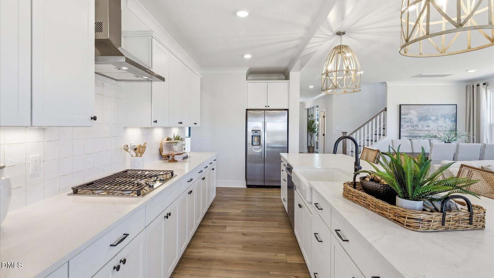 Modern kitchen with white cabinets, stainless steel appliances, wooden floor, two pendant lights, a gas stove, and a basket of potted plants on a white island. Living area and staircase are visible in the background.