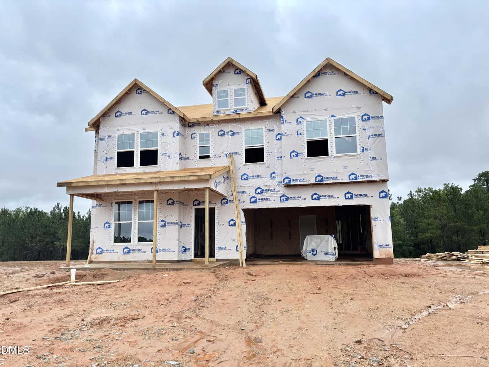 The two-story house at 117 Thompson Pond Way, Franklinton is under construction, wrapped in weatherproof sheeting with window openings and an unfinished porch. The ground is bare dirt, and a forested area is visible in the background under a cloudy sky.