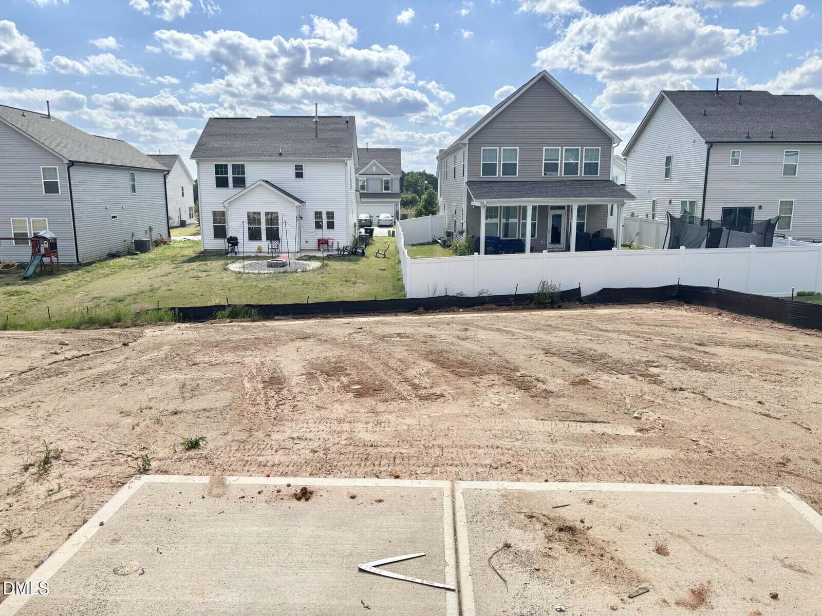 A cleared dirt lot sits in front of two gray and white suburban houses with backyards, separated by a white fence, under a partly cloudy sky.