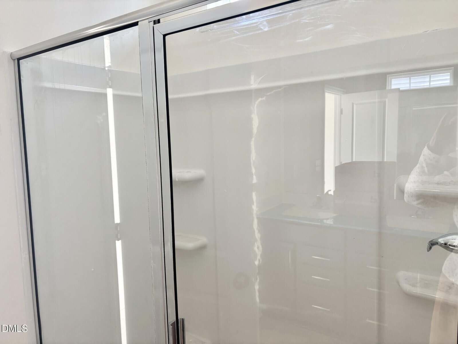 A glass shower door with metal framing, showing some streaks and reflections. In the reflection, a bathroom vanity with drawers, double sinks, and an open door are visible.