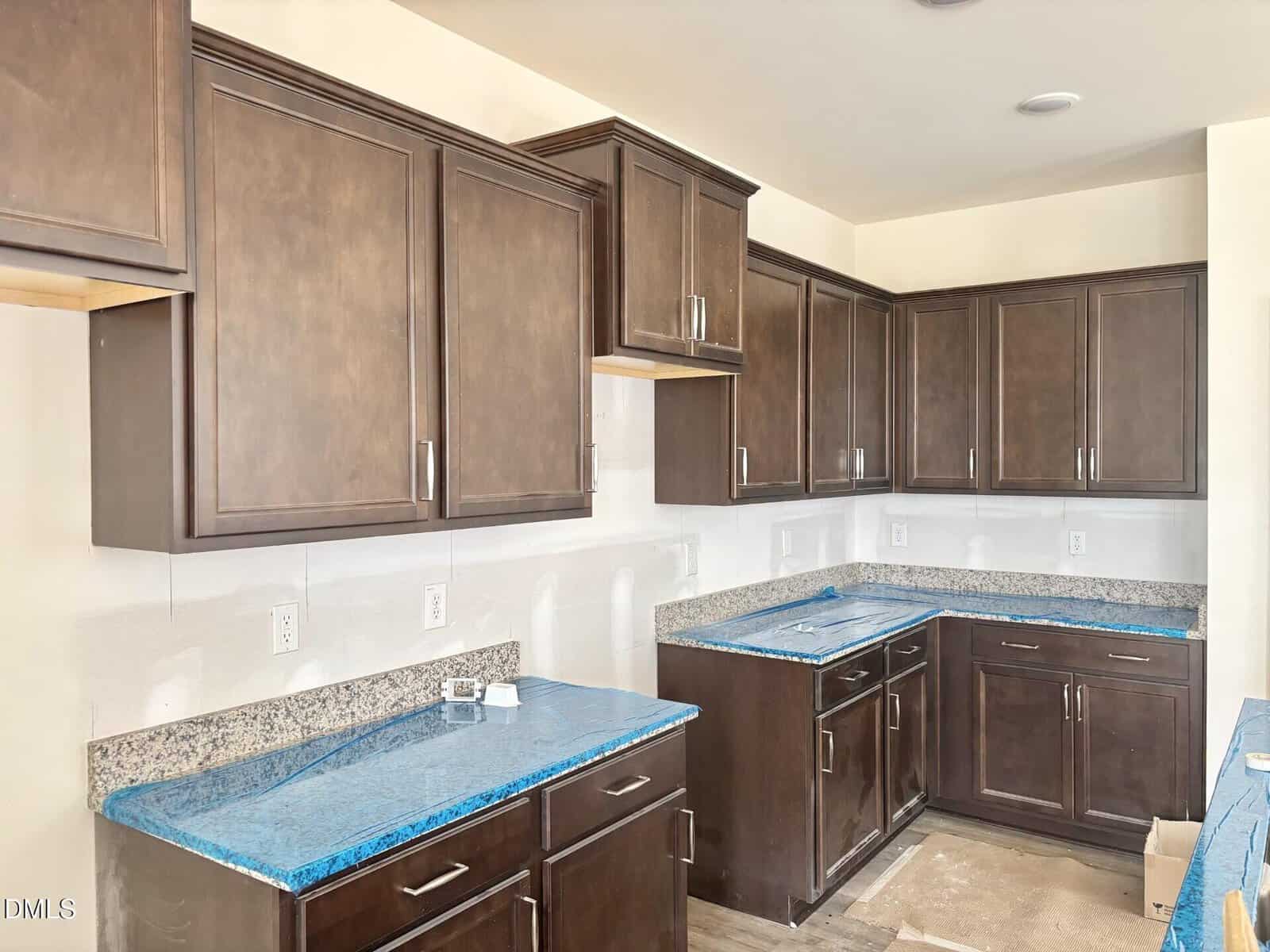 A kitchen under construction with dark wood cabinets, light granite countertops covered in blue protective film, and white walls. The floor is unfinished and there are no appliances installed yet.