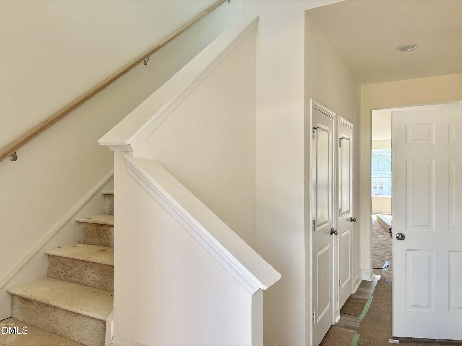 A staircase with unfinished wooden steps and a simple handrail leads up from a hallway with beige walls. Nearby are two closed closet doors and an open white door revealing a sunlit room.