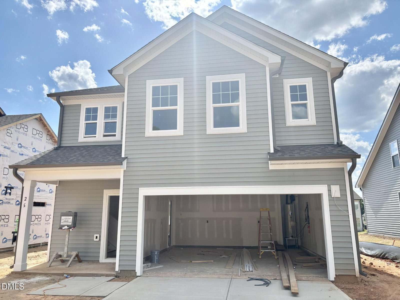 A two-story gray house under construction with three garage doors open, exposed framing and construction materials inside, and neighboring homes in progress visible under a partly cloudy sky.