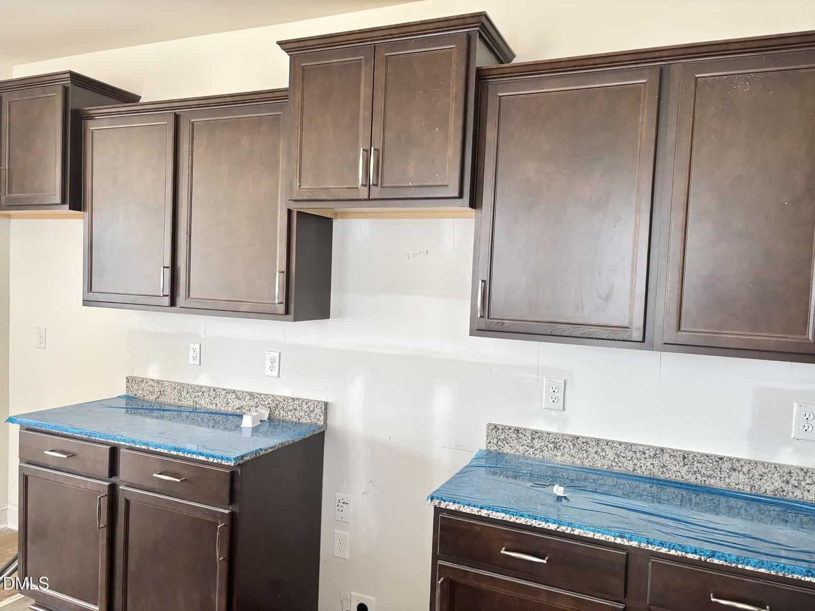 Kitchen with dark brown cabinets, granite countertops covered in protective blue plastic, white walls, electrical outlets, and an empty space for a stove or microwave between cabinets.