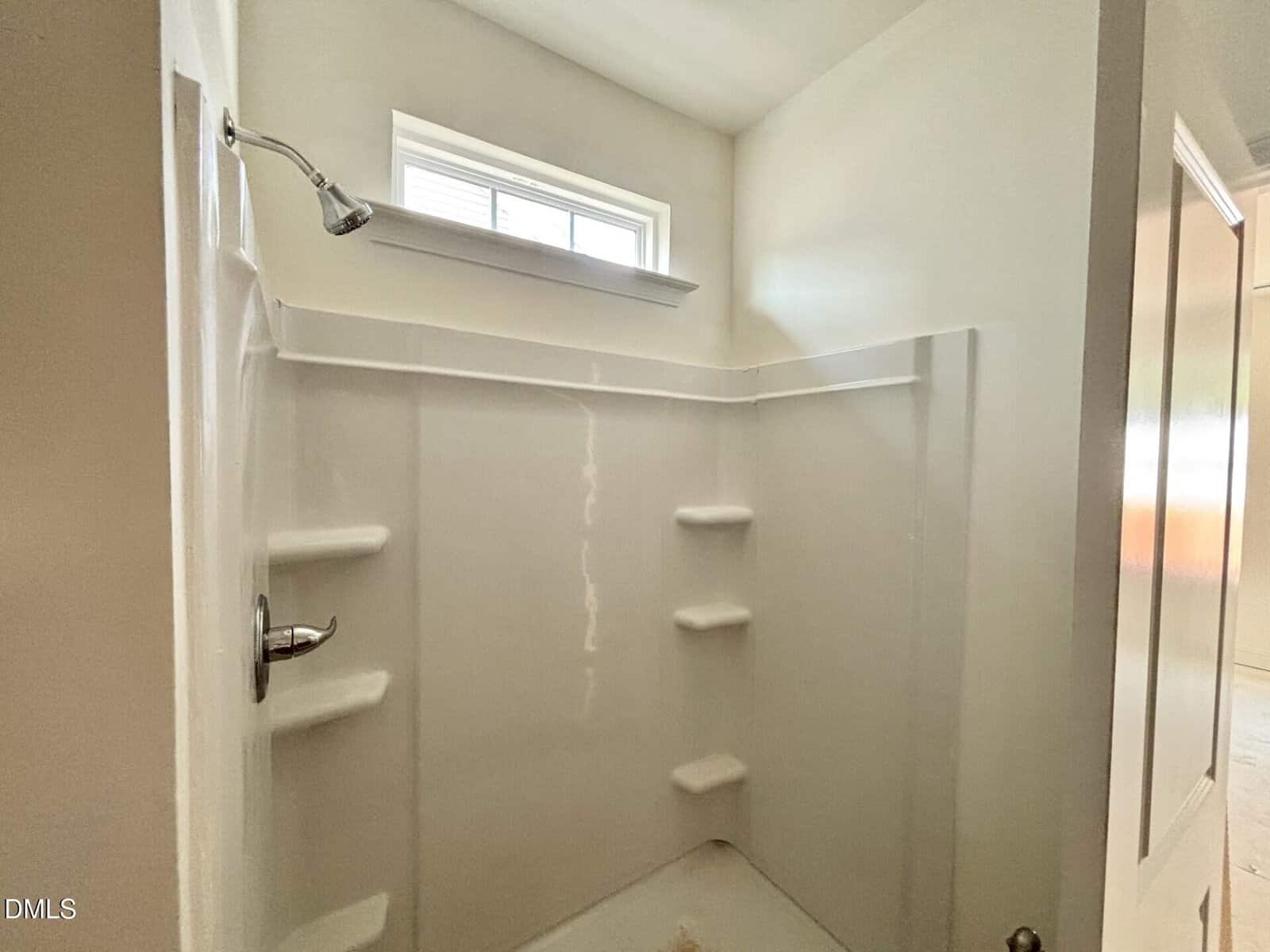 A white shower stall with built-in corner shelves, a silver showerhead, and a small rectangular window above letting in natural light. The shower is surrounded by smooth white walls and an open door is visible.