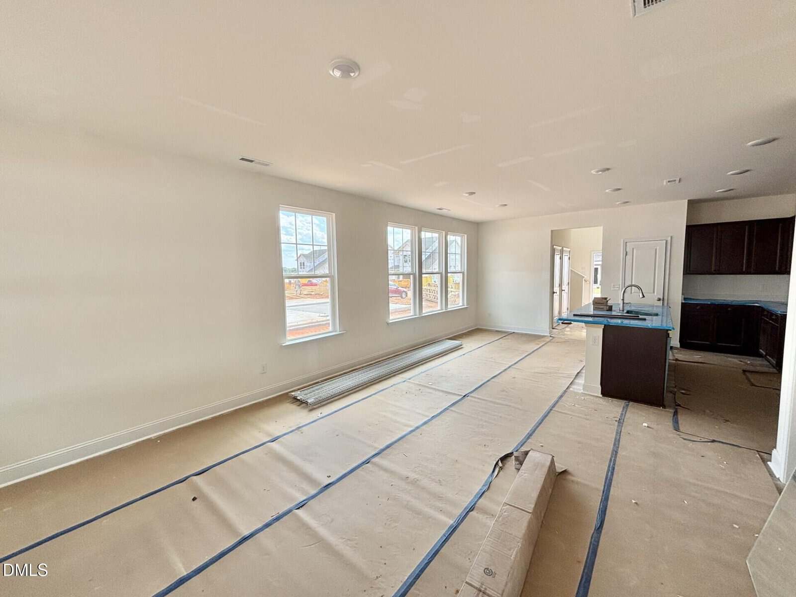 Unfurnished, newly constructed open-plan living and dining area with large windows, covered floors, and an adjacent kitchen featuring dark cabinets and a center island. Construction materials are visible on the floor.