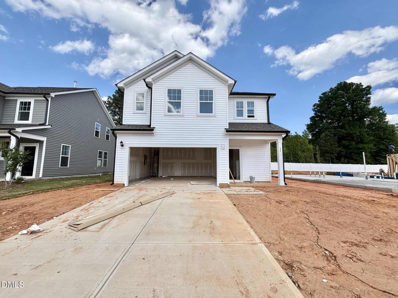 A newly constructed two-story white house with an attached garage, unfinished driveway, and bare dirt yard. The sky is partly cloudy, and construction materials are visible near the house.