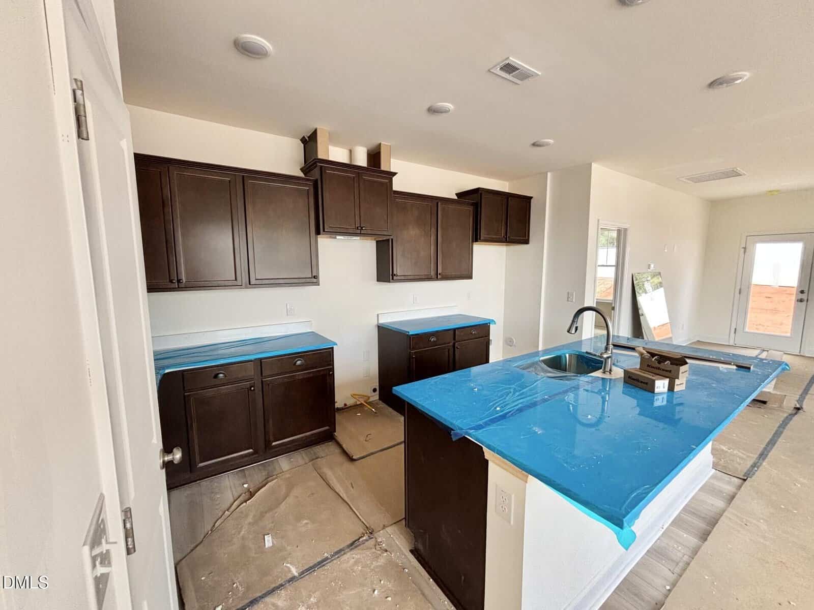 A modern kitchen under construction with dark wood cabinets, blue protective plastic covering the countertops, an island with a sink, and unfinished floors. Light streams in through a door and window in the background.