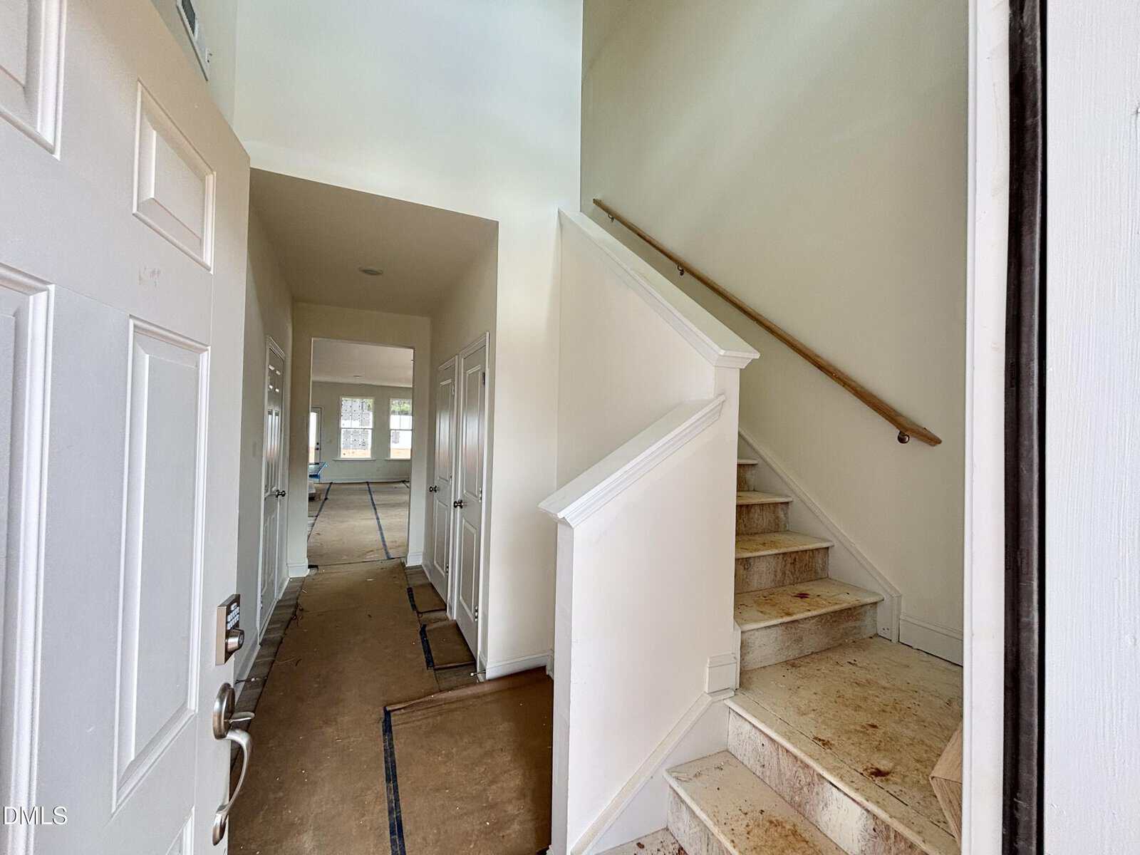 Open white front door leading to a hallway with a staircase on the right. The stairs are partially carpeted and slightly stained. Down the hall, there are doors on the left and a bright room at the end.